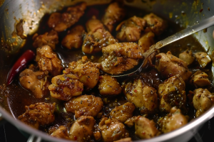 Chicken pieces sizzling in a hot frying pan with aromatic spices and herbs during Pallipalayam Chicken Gravy prep